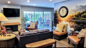 Ein Wohnzimmer mit einer Couch, Sesseln, einem Couchtisch, einer Lampe, einem Kamin und einem großen Fenster mit Blick auf Gartenmöbel.