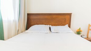 Un lit bien fait, avec du linge de lit blanc et une tête de lit en bois, une petite table sur laquelle est posé un vase de fleurs, et une chaise à droite.