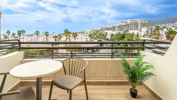 Un balcon avec une table ronde et deux chaises, un pot de fleurs et une vue sur un paysage urbain côtier, avec des palmiers et des bâtiments.