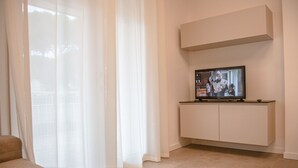 A modern living room with a flat-screen TV on a white stand, beige curtains, and a beige sofa.