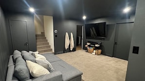 A modern living room with a grey sectional sofa, a white round ottoman, and a flat-screen TV mounted on the wall.