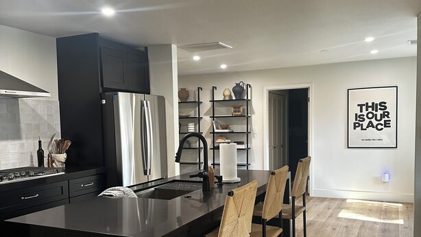 A modern kitchen with a central island, black cabinetry, and a black countertop. There are four bar stools with woven seats. A black refrigerator and a stove are visible. The kitchen has a white backsplash and a ceiling with recessed lighting. A framed quote on the wall reads "THIS IS OUR PLACE."