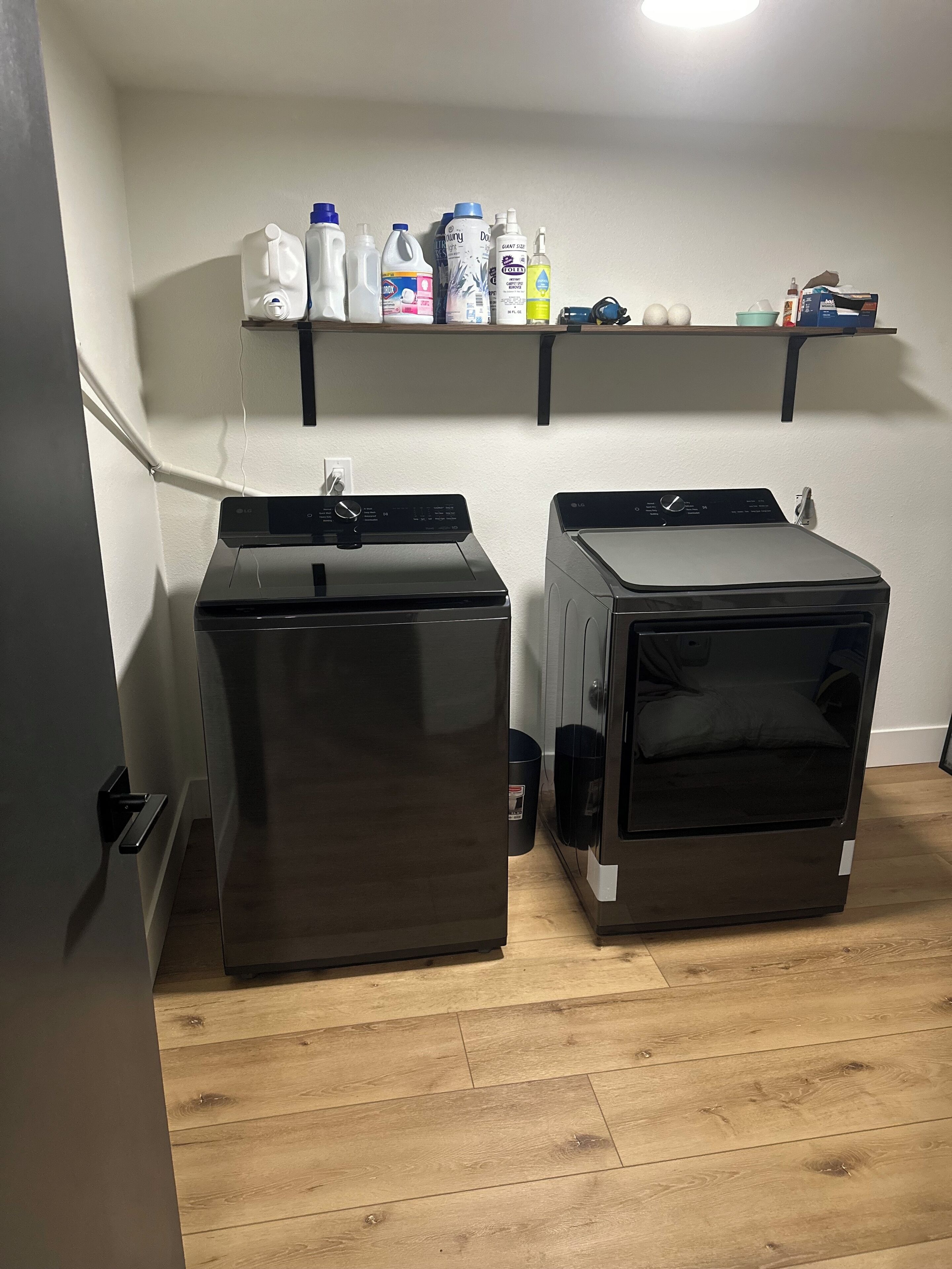 Un cuarto de lavandería con dos lavadoras de carga frontal y un estante superior con diversos productos de limpieza.