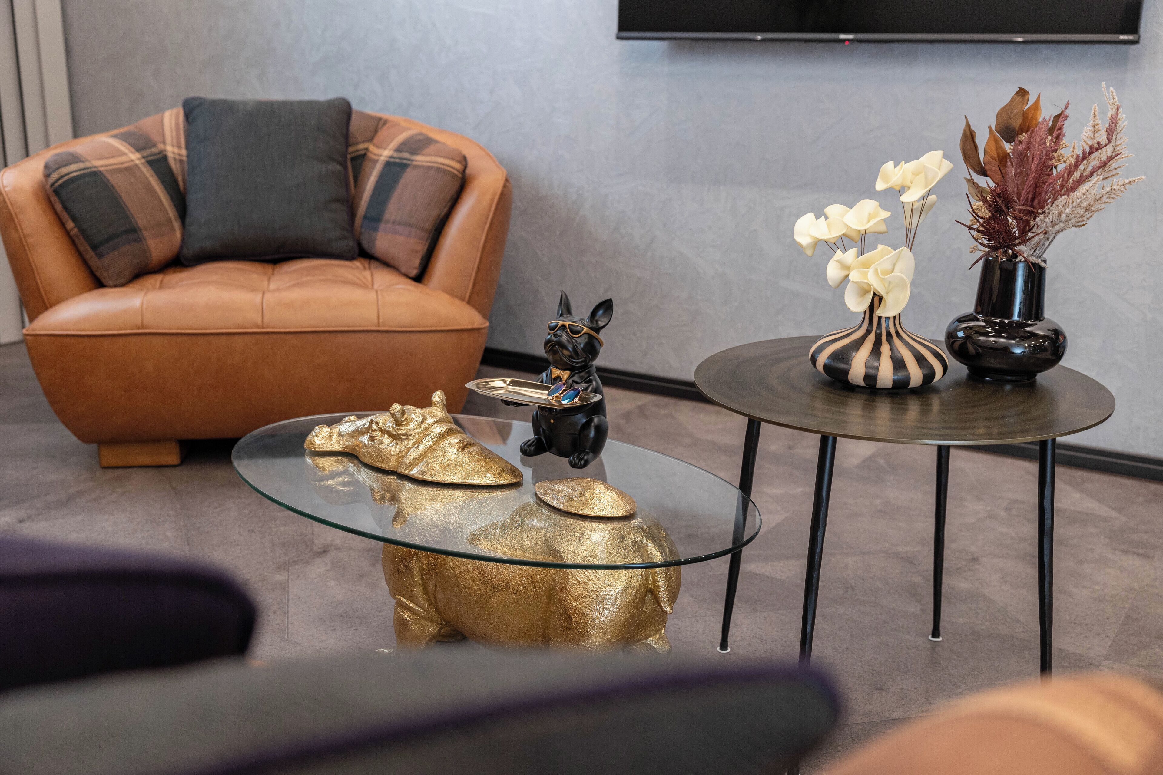A modern living room with a brown leather sofa, a glass coffee table with gold figurines, and two side tables with decorative vases.