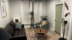 Een woonkamer met een bruine leren bank, een groene fauteuil, een ronde salontafel en een kamerbreed tapijt. Op een klein tafeltje staat een potplant, er is een wandventilator en een deur met een decoratief object erboven.