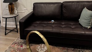 Een woonkamer met een donkerbruine leren bank, een lijst met een botanische tekening aan de muur, een bijzettafeltje met een lamp, en een kamerkleed met een patroon.