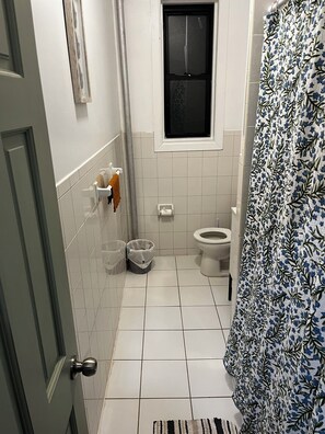 Un baño pequeño con azulejos blancos, inodoro, ventana y cortina de ducha.