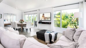 Een moderne woonkamer met een grote televisie, comfortabele zithoek, een eethoek en grote ramen met uitzicht op groen.