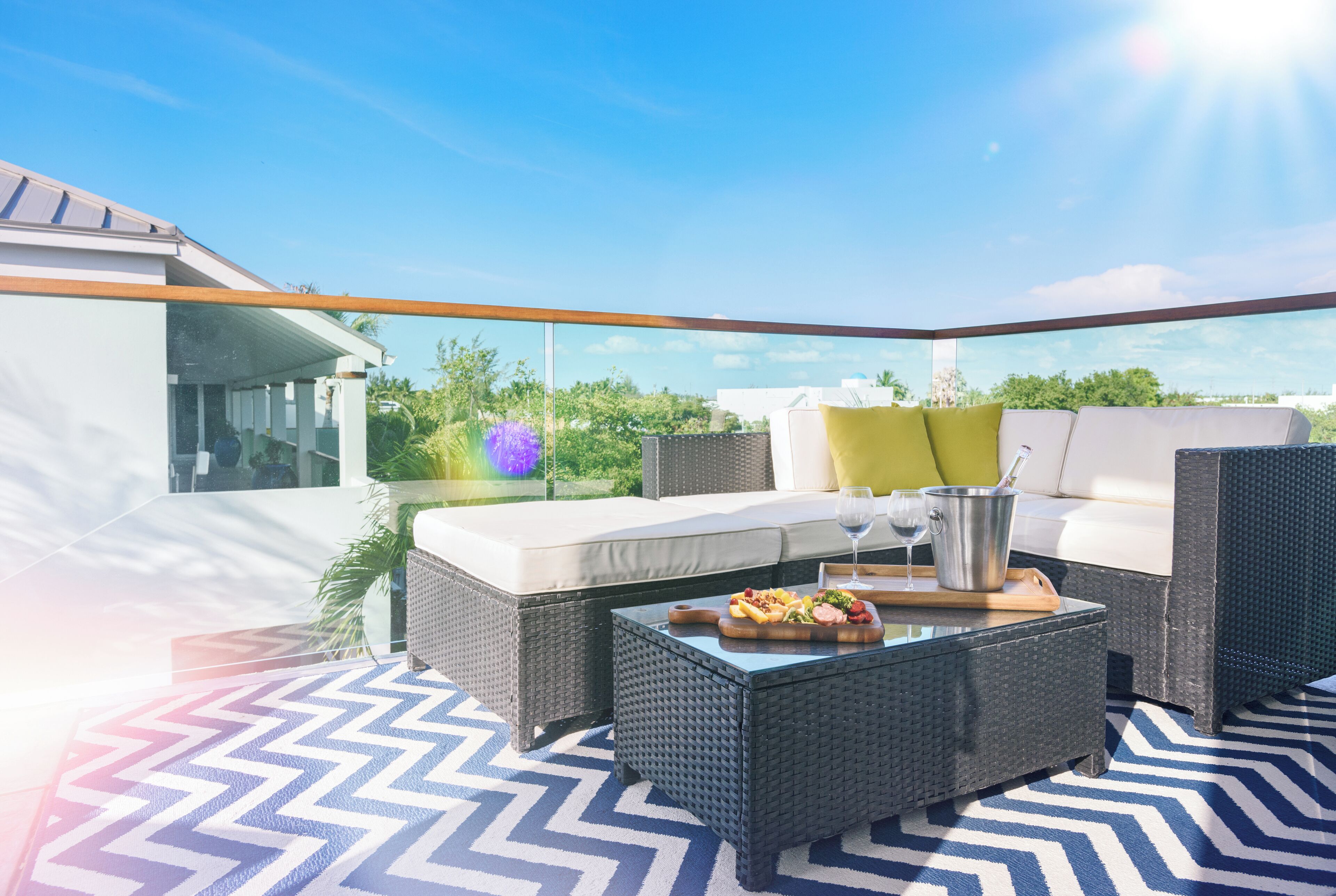 An outdoor patio with wicker furniture, a glass table with food, and a view of greenery and buildings.