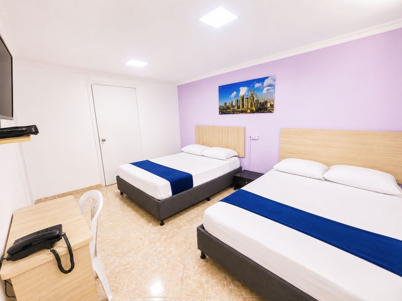 Hotellihuone, jossa on kaksi sänkyä, työpöytä puhelimineen ja seinällä kaupunkimaisemaa esittävä taulu.