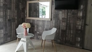 Quarto moderno com um espelho grande, televisão de tela plana, duas cadeiras brancas e uma mesa redonda pequena com um vaso de flores.