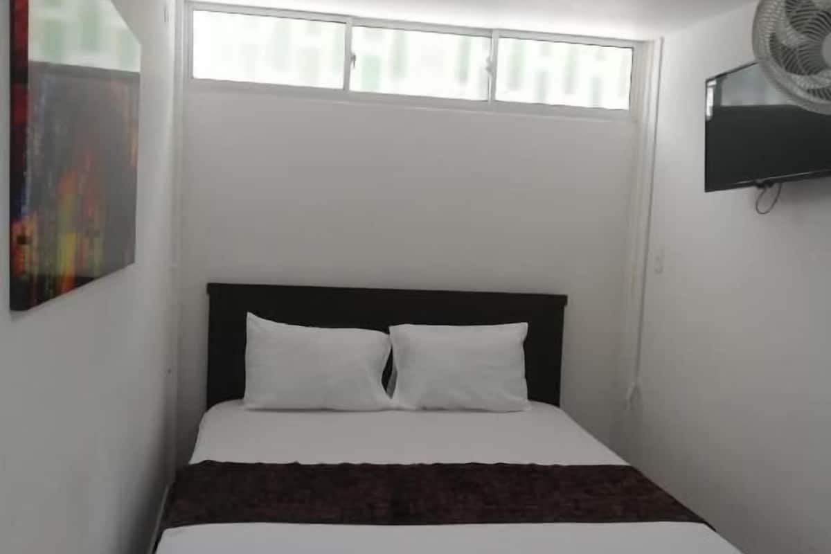 A small, neatly made bed with white bedding and a dark headboard, positioned under a window with blinds.