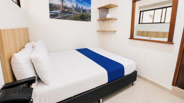 A hotel room with a bed, a mounted television, a window with blinds, a painting on the wall, and a wooden shelf unit. - Sol de Estadio (Medellín)