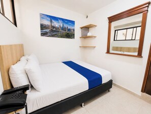 A hotel room with a bed, a mounted television, a window with blinds, a painting on the wall, and a wooden shelf unit.