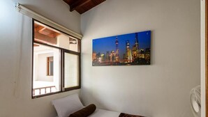 Quarto de hotel com cama, janela com persianas, pintura da paisagem urbana na parede e um ventilador.