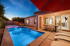 A swimming pool with a patio area, a dining table with chairs, and a modern kitchen visible through glass doors. - Pool + spa home in gated resort (Indio)