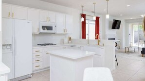 A modern kitchen with white cabinets, a central island, and a dining area with a TV and red curtains. - Stylish Indio Sanctuary with Heated Pool (Indio)
