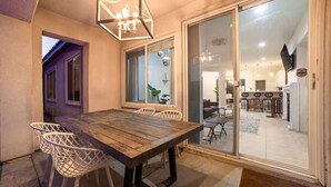 A dining area with a wooden table and white chairs, a chandelier, and a sliding glass door leading to a kitchen and living space. - Stylish Indio Sanctuary with Heated Pool (Indio)