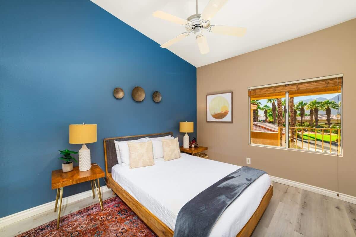 A bedroom with a bed, two bedside tables with lamps, a ceiling fan, a window with a view of palm trees, and a patterned rug.