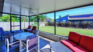 A covered patio with a round table and chairs, a red sofa, and a view of a blue house and a wooden fence.