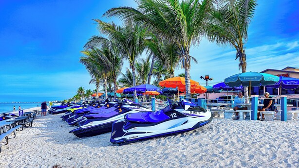 A beach with jet skis lined up, palm trees, and beach umbrellas.
