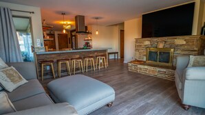 A modern living room with a stone fireplace, a bar area with stools, and a large TV mounted on the wall.