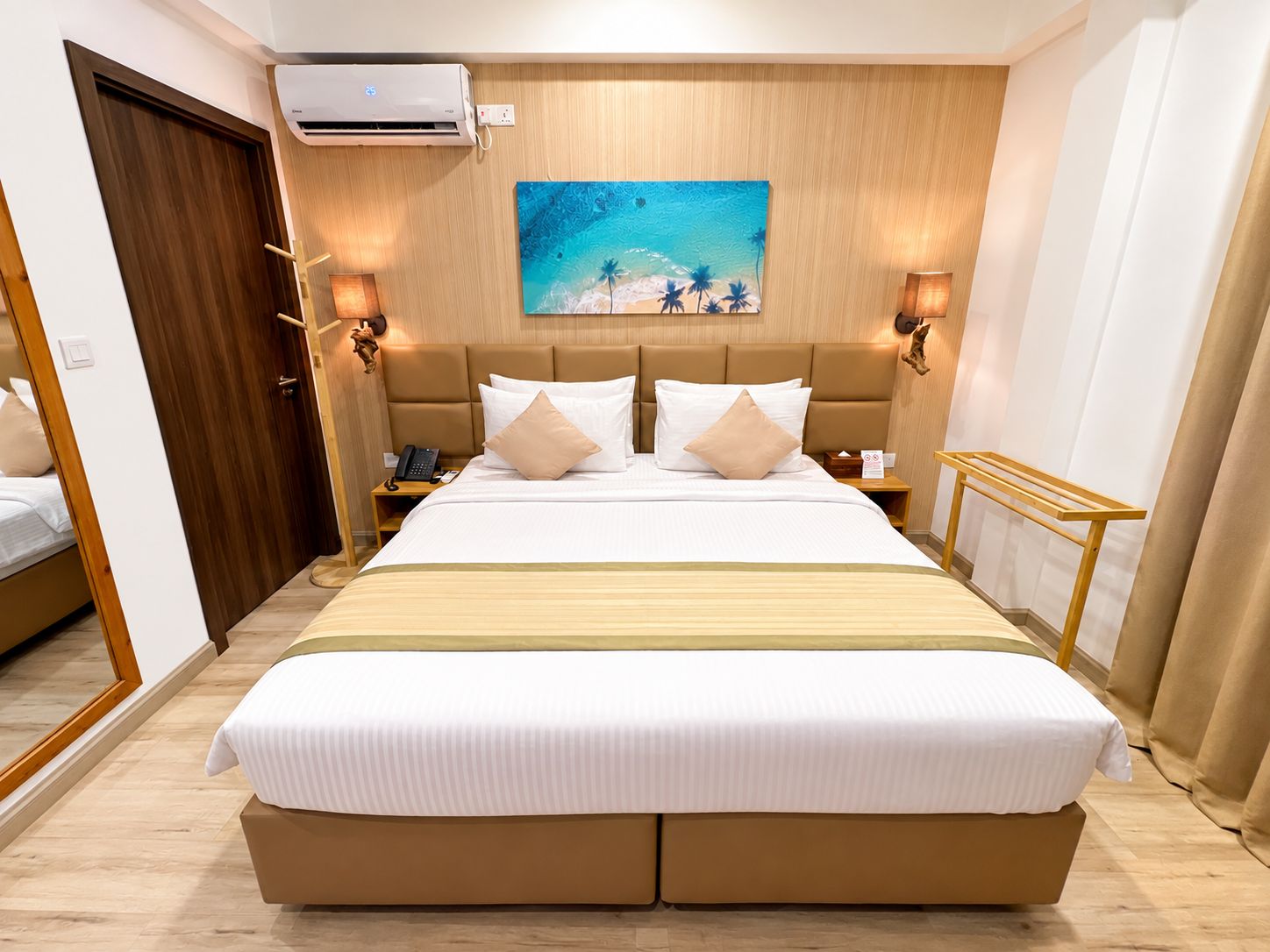 Une chambre d’hôtel avec un grand lit, un tableau représentant une scène de plage, un climatiseur fixé au mur et des lampes de chevet.