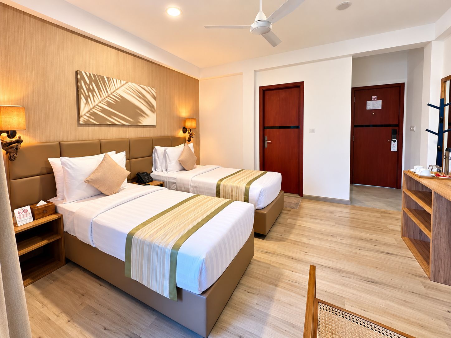 Quarto de hotel com duas camas, cabeceira de madeira, luminárias de cabeceira, ventilador de teto e piso de madeira.