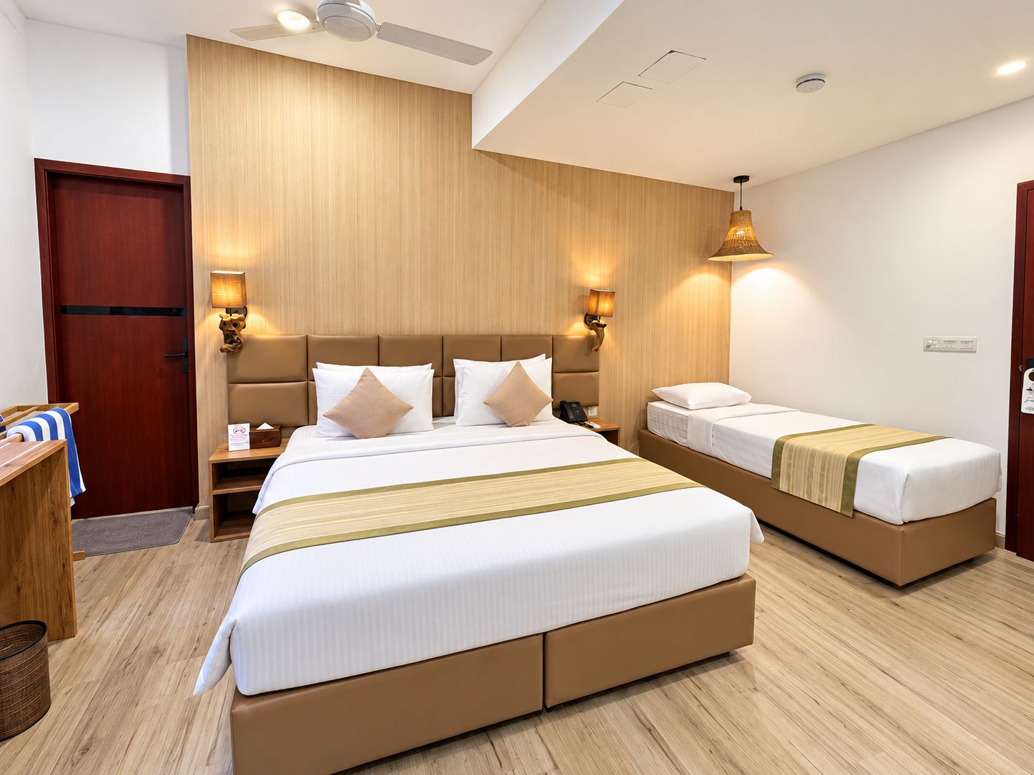 A hotel room with two beds, a wooden headboard, bedside lamps, a desk, and a ceiling fan.