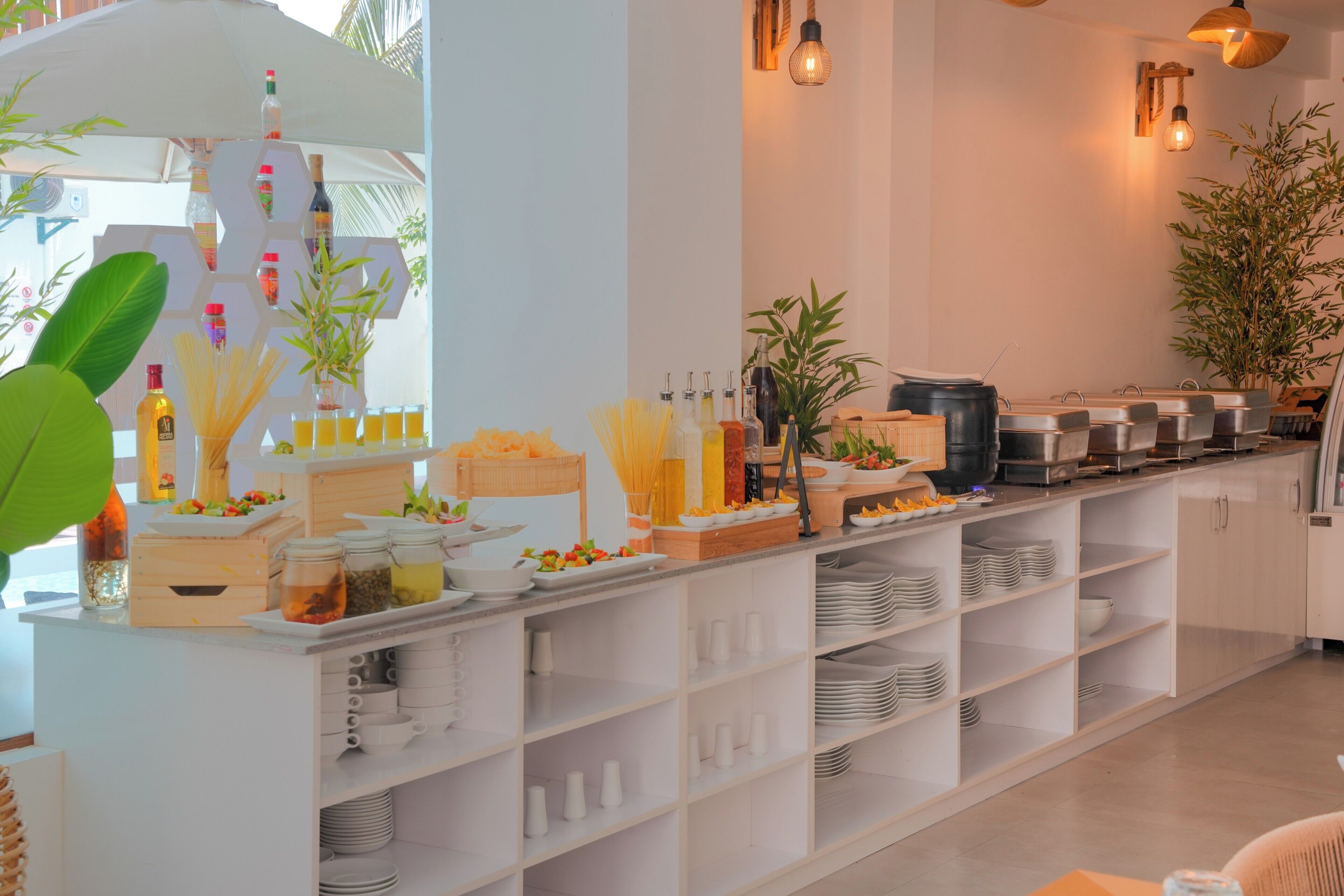 A buffet setup with various food items and beverages, including pasta, fruits, and condiments, arranged on a white counter with shelves below.