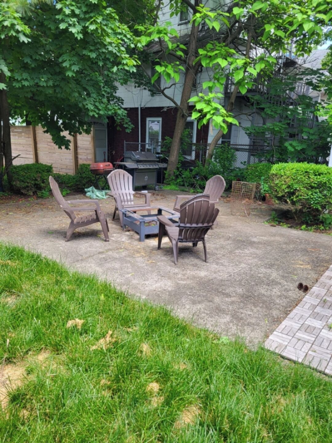 An outdoor seating area with Adirondack chairs and a table, surrounded by greenery and a barbecue grill.