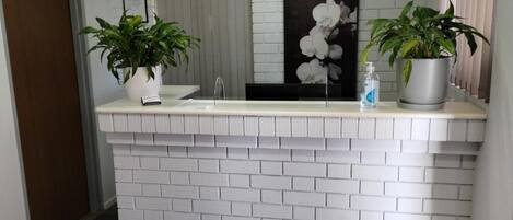 A reception area with a white brick wall, a white countertop, two potted plants, a framed picture, and a wall-mounted air conditioner.