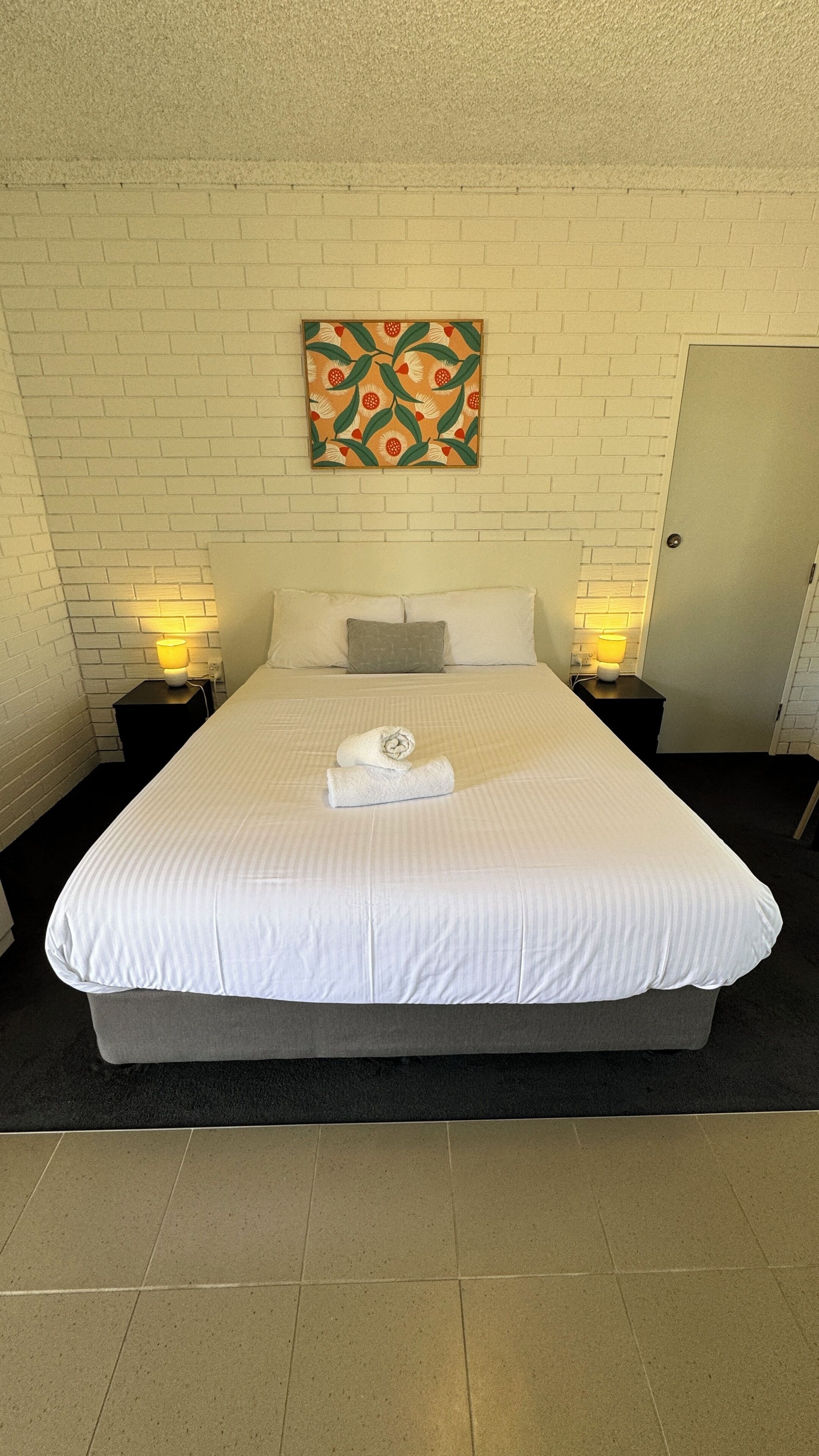 A neatly made bed with white linens, two bedside tables with lamps, a wall-mounted artwork, and a door to the right.