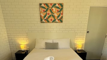 A neatly made bed with white linens, two bedside tables with lamps, a wall-mounted artwork, and a door to the right.