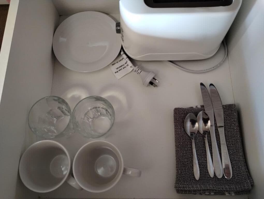 Una mesa blanca con un tostador, dos vasos vacíos, dos tazas vacías y un servilleta con cuchara y cuchillo.