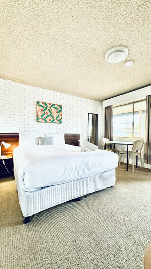 A hotel room with a large bed, a desk, and a chair. There is a painting on the wall and a window with curtains. - Junction Motel Wagga (Ashmont)
