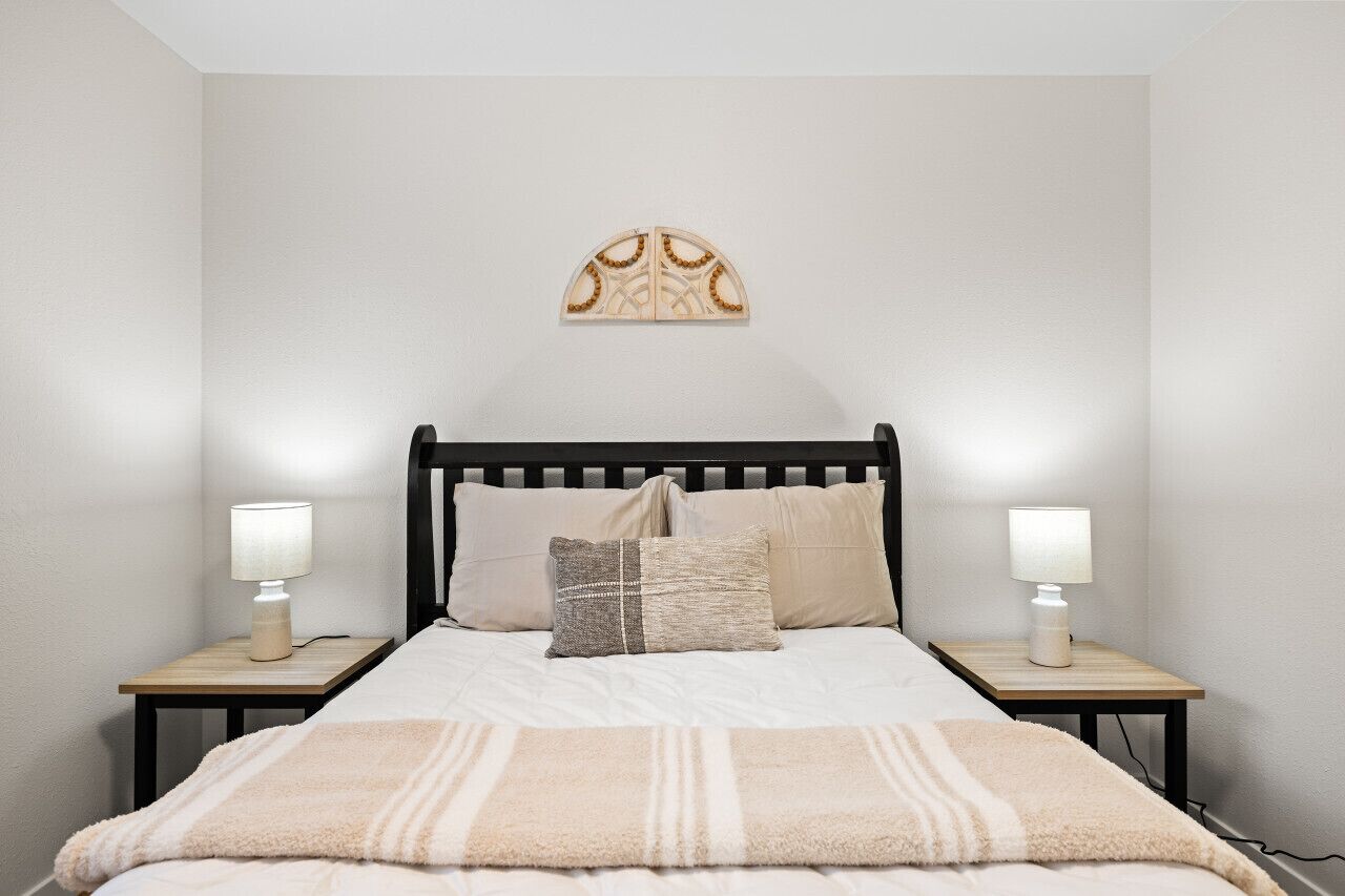 Quarto com cama, duas mesas de cabeceira, duas luminárias, um detalhe decorativo na parede e um cobertor.