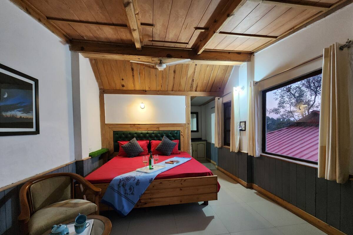 Ein Schlafzimmer mit Holzboden, einem Bett mit roter Bettwäsche, einem Nachttisch mit darauf liegenden Gegenständen und einem Fenster mit Blick auf ein Gebäude mit rotem Dach.