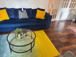 A dark blue sofa with yellow and gray cushions, a glass coffee table with a plant, and a yellow rug on a wooden floor.