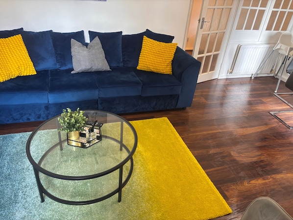A dark blue sofa with yellow and gray cushions, a glass coffee table with a plant, and a yellow rug on a wooden floor.
