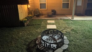 Ein Garten mit Feuerstelle, Lichterkette, Holzzaun und einem Haus mit Fenstern.