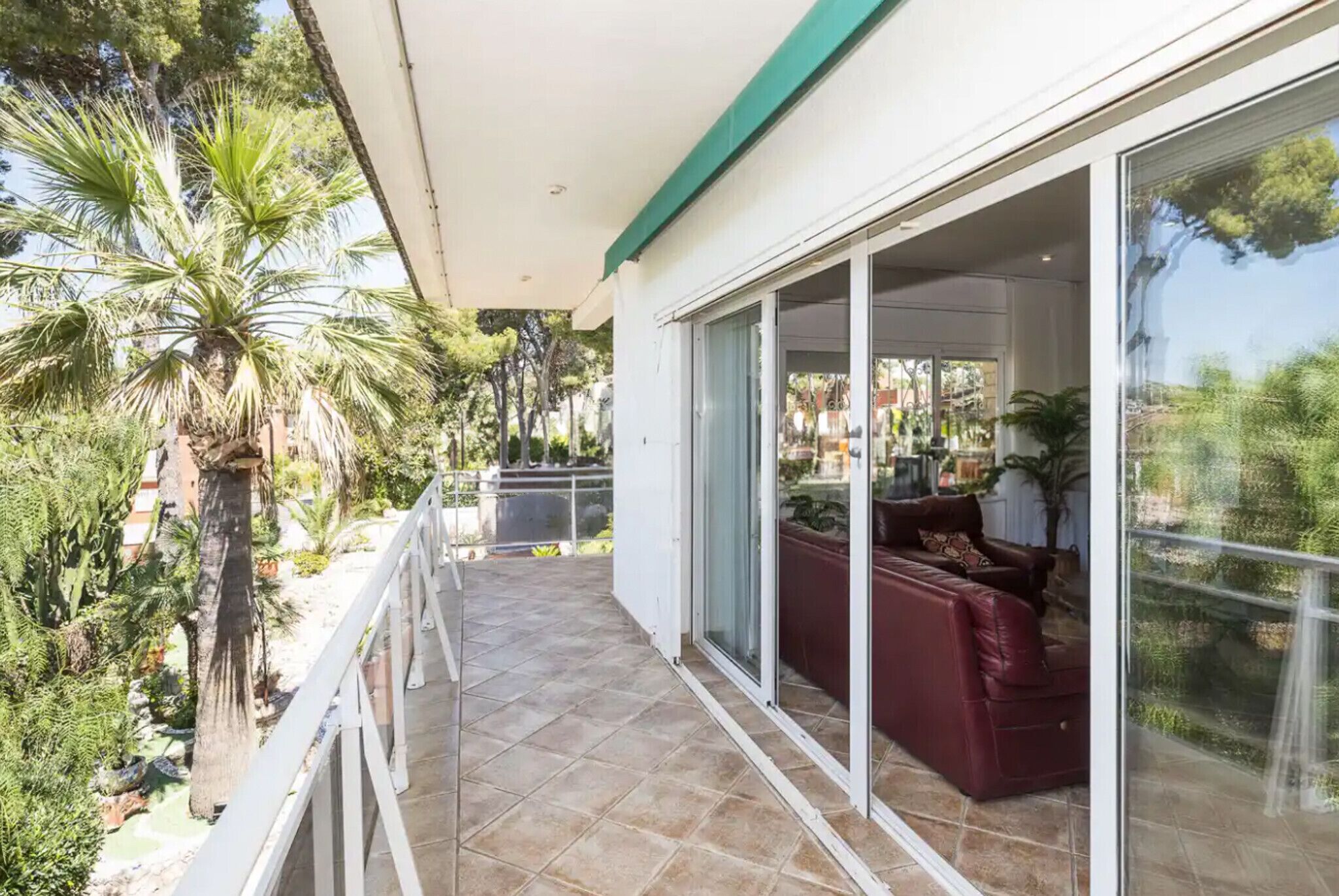 Ein Balkon mit einer roten Couch, einer Glastür und Blick auf einen Garten mit Palmen.
