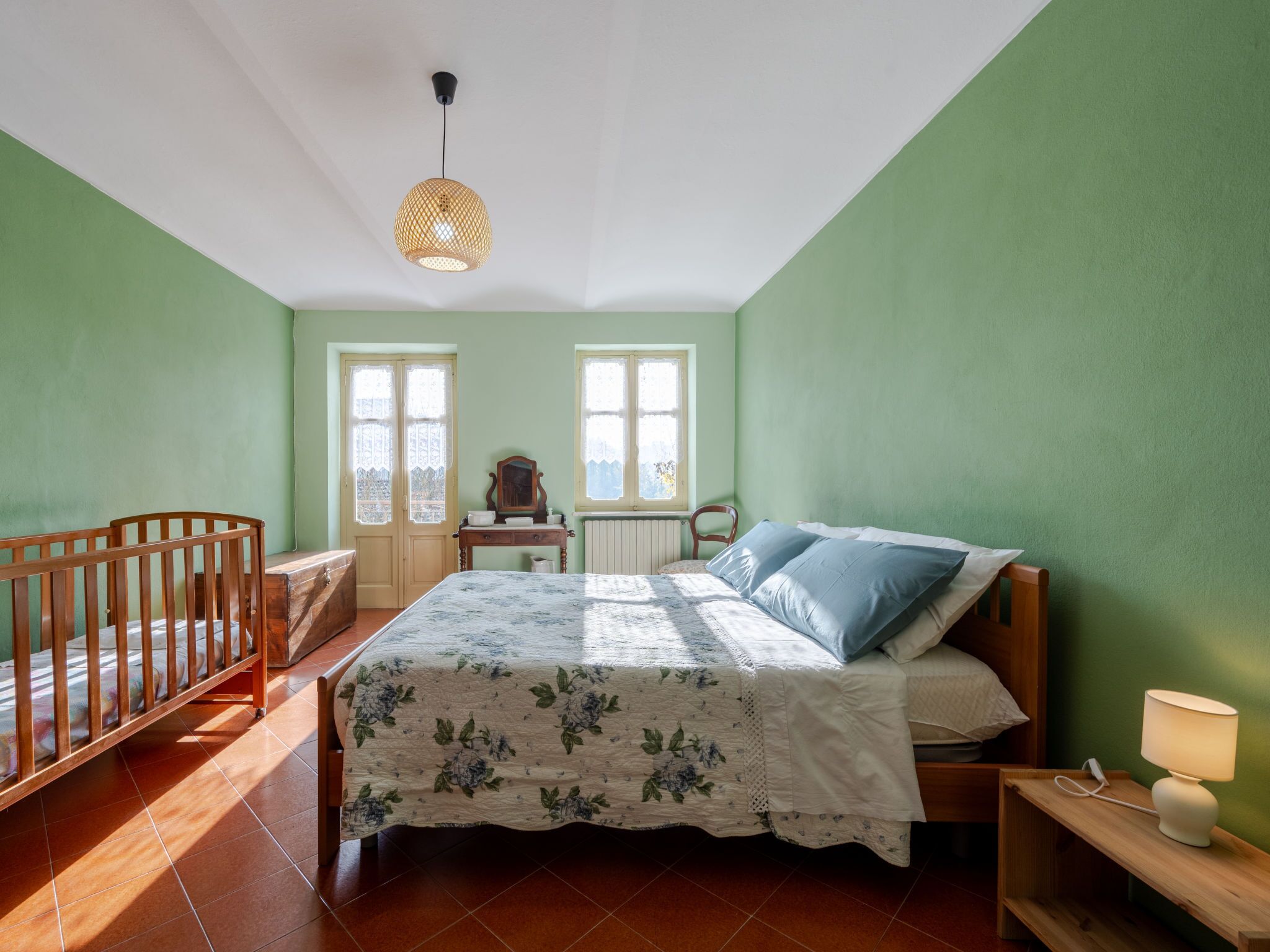 Ein Schlafzimmer mit einem Bett, einem Kinderbett, einer Kommode, einem Nachttisch, einer Lampe und zwei Fenstern.