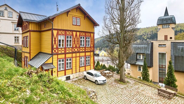 Un bâtiment jaune avec une voiture garée devant, entouré de verdure et bordé d’un chemin pavé.