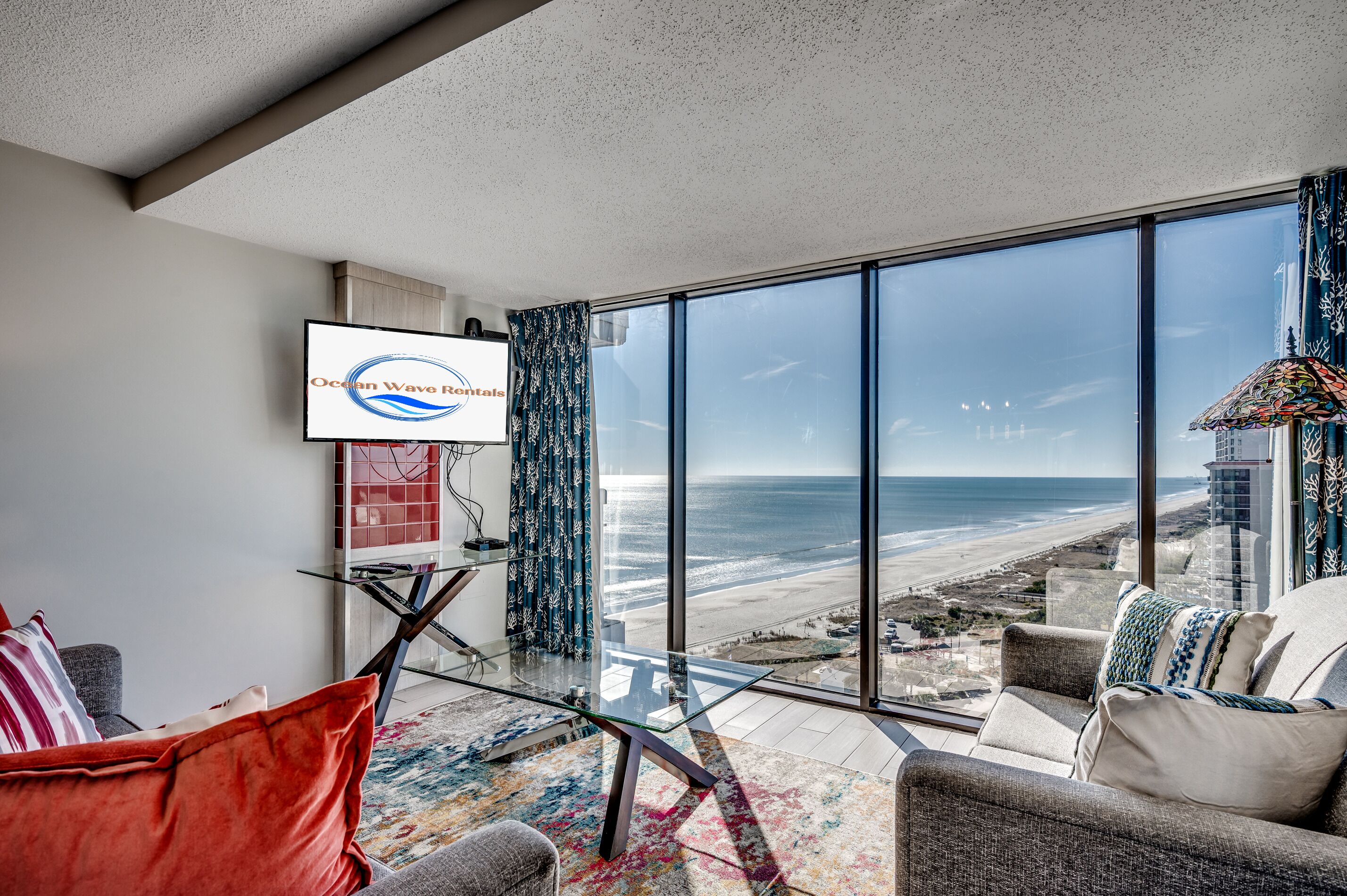 A modern living room with a large glass table, a sofa, and a TV displaying 'Ocean Wave Rentals'.