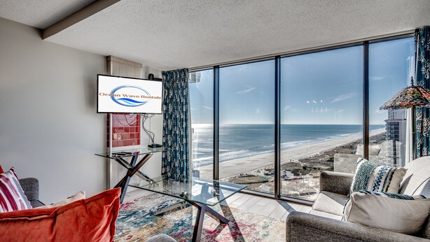 A modern living room with a large glass table, a sofa, and a TV displaying 'Ocean Wave Rentals'. - Sweeping Ocean View from Each Bdr and Living Room, Safe and Quiet Neighborhood  (Myrtle Beach)