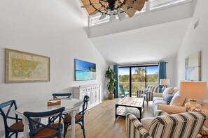 A spacious living area with a dining table, chairs, a sofa, and a coffee table. There is a large window with a view of the outdoors. - Osprey's Nest | Luxury Sea Palms Golf Condo St. Simons Island (Saint Simons Island)