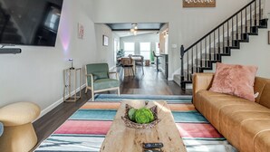 Een ruime woonkamer met een grote televisie, een houten salontafel met afstandsbediening, een groene fauteuil en een bruine bank met een roze kussen. Een trap leidt naar een hoger gelegen verdieping.
