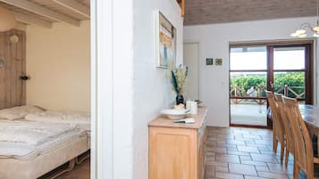 Quarto com cama, cômoda de madeira, mesinha com um vaso e porta que dá para uma varanda com vista para o verde.