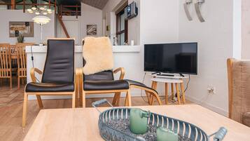 Uma sala de estar moderna com uma mesa de jantar de madeira, duas poltronas de couro, uma TV sobre suporte e uma área de cozinha ao fundo.
